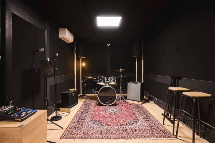 Rehearsal room seen in its entirety, with a complete drum kit in the center, a bass amp, a guitar amp and a mixer along the walls, and a Persian rug on the floor. | © Plug The Jack