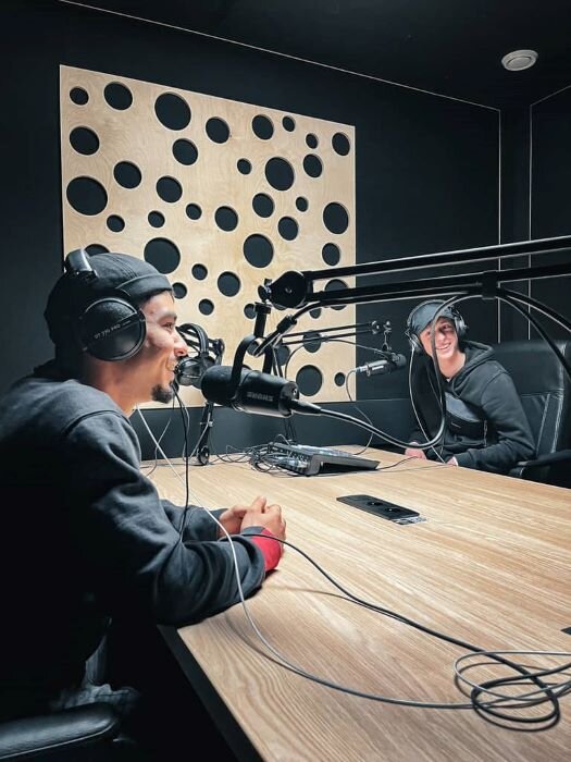 Podcast studio with two cheerful hosts around a table equipped with microphones. | © Plug The Jack