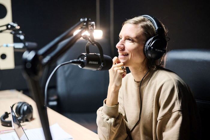 Vrouw voor een Shure MV7 podcastmicrofoon, pratend met koptelefoon op. | © Plug The Jack
