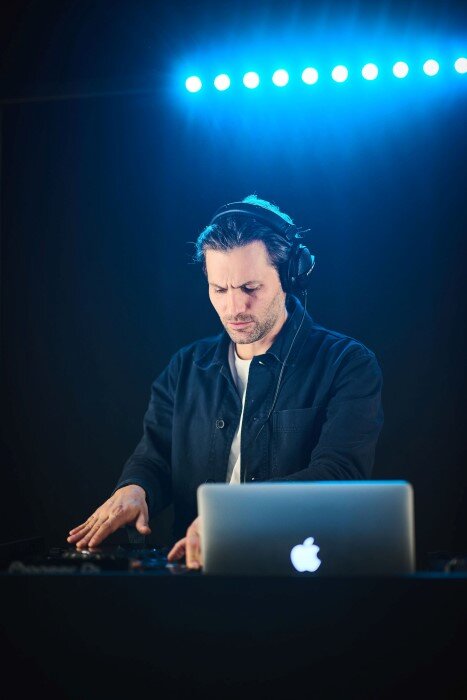 Man in DJ-studiokamer met CDJ-3000, koptelefoon en blauwe spotlights aan het mixen. | © Plug The Jack